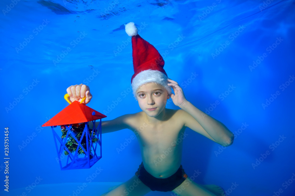 A little boy swims underwater in the red hat of Santa Claus and holds a gift in his hand. Portrait. Landscape orientation A little boy swims underwater in the red hat of Santa Claus and holds a gift in his hand. Portrait. Landscape orientation