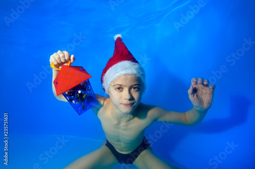 Portrait of a little boy who posing underwater at the bottom of the pool in a red hat of Santa Claus with a toy in his hand. He looks at the camera and smiles. Landscape orientation