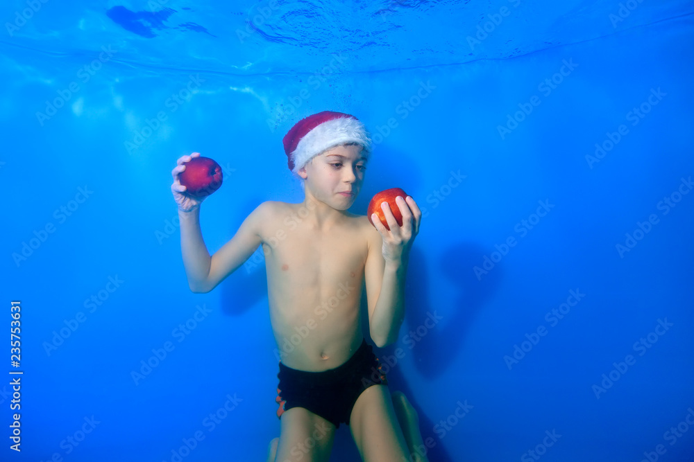The little boy looks at the red apple in his hand with surprise. He swims underwater on a blue background in Santa's hat. Portrait. Concept. Horizontal orientation