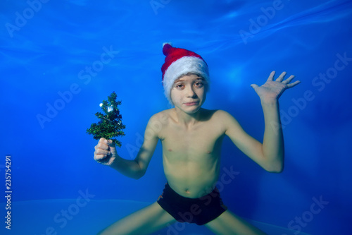 Sad little boy posing under water at the bottom of the pool in a Santa Claus hat with a Christmas tree in his hand on a blue background and looks at the camera. Portrait. Horizontal orientation