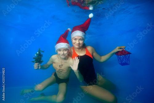 A little boy with his mother posing under water at the bottom of the pool in a Santa hat, holding Christmas toys in their hands, look at the camera and smile. Portrait. Concept