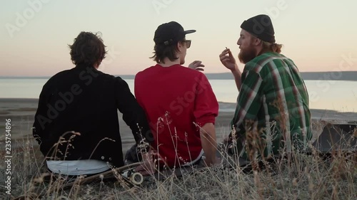 Back view of three men smoke weed at sunset sit at sea coast rapid slow motion