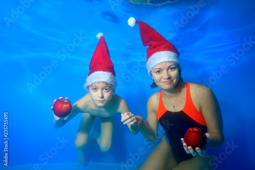 Mother and child posing together underwater on blue background, holding in hands red apples in a hat of Santa Claus smiling and looking at the camera. Portrait. Landscape orientation