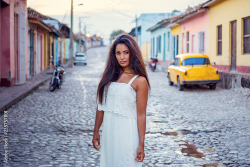 Photography Beautiful dark skinned young lady in white dress standing in front of an old classic car and a motorcycle in the old streets of Trinidad in Cuba