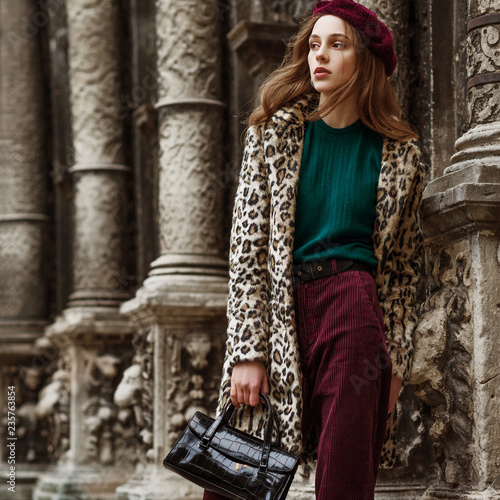 Outdoor fashion portrait of woman wearing trendy animal, leopard print faux fur coat, beret, sweater, corduroy trousers, holding reptile skin textured bag, posing in street of city. Copy, empty space
