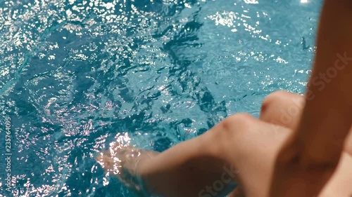 Close-up legs of tanned young woman relaxing and having good time sitting on poolside high angle barefoot female sunbathing playing feet in pure water at outdoor swimming pool slow motion top view