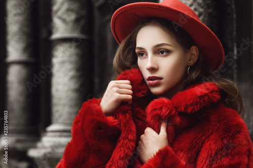 Outdoor close up fashion portrait of young beautiful confident woman wearing trendy orange faux fur coat, hat, silvery hoop earrings, posing in street of european city. Copy, empty space for text