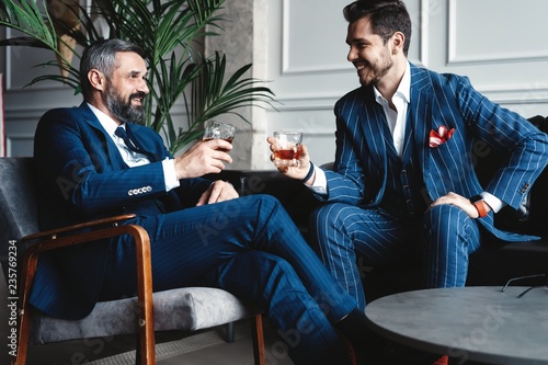 Relaxing. Full length of two young handsome men in suits holding glasses and looking at each other while resting indoors. photo