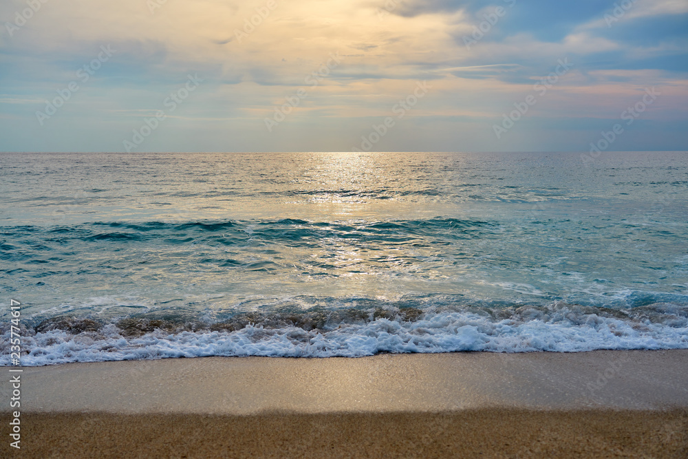 Fototapeta premium Sunset at the tropical beach, sun behind clouds reflects on water and waves with foam hitting sand.