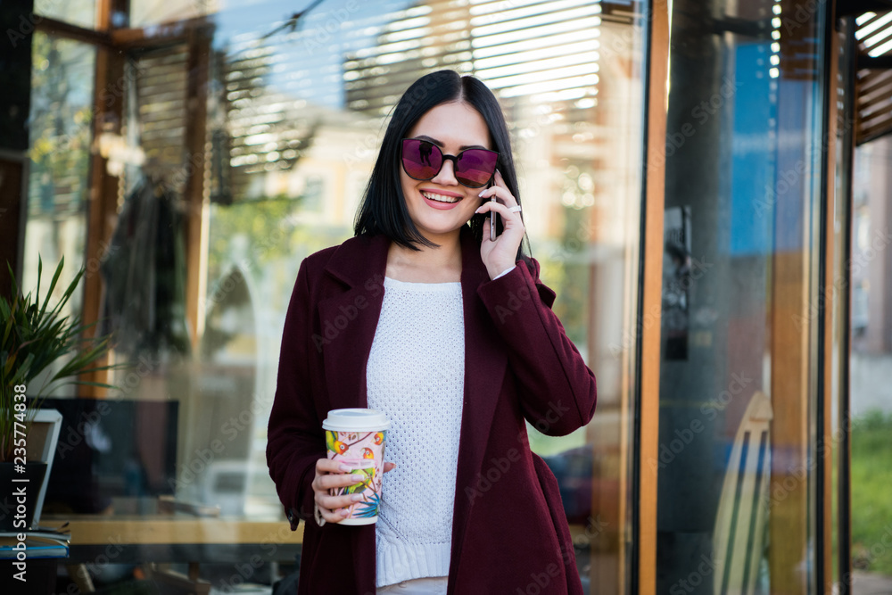 Fototapeta premium Portrait pretty girl with dark hair in Burgundy fur coat of artificial fur. She looks excited and talking on a mobile phone. City lifestyle.