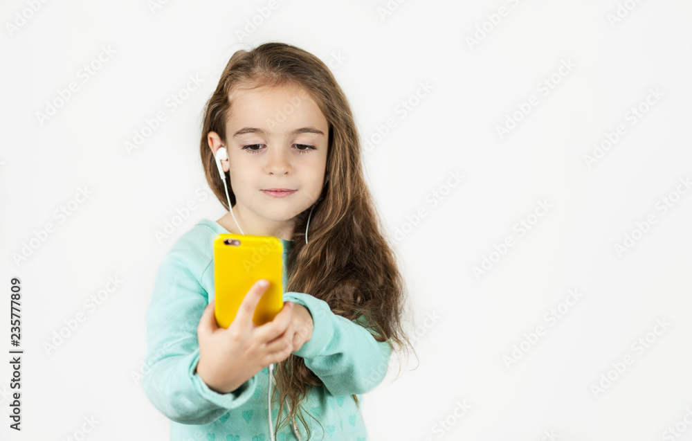 Bright beautiful little girl in headphones with the phone emotionally posing for a selfie on an isolated background. Bright beautiful little girl in headphones with the phone emotionally posing for a selfie on an isolated background.