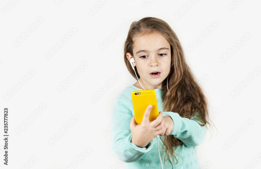 Bright beautiful little girl in headphones with the phone emotionally posing for a selfie on an isolated background. Bright beautiful little girl in headphones with the phone emotionally posing for a selfie on an isolated background.