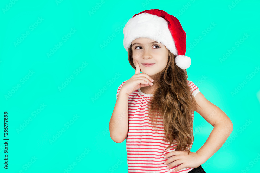 Little bright beautiful girl emotionally posing in Santa hat on an isolated turquoise background Little bright beautiful girl emotionally posing in Santa hat on an isolated turquoise background