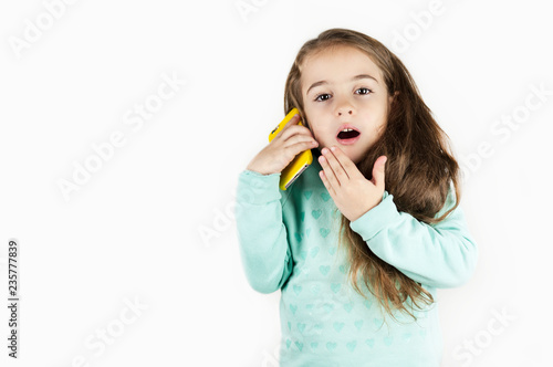 Bright beautiful little girl in headphones with the phone is emotionally surprised on an isolated background.