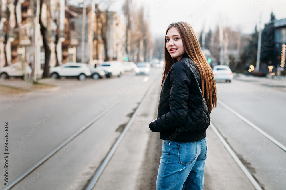 Portrait from back of elegant fashion girl long hair walking on on city background. She has black leather jacket and jeans. She is smiling to camera.