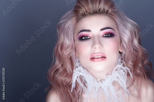 Closeup portrait of beautiful young model with creative colorful make up and wavy hairstyle wearing stylish white feather collar. Isolated on grey background. Text space. Studio shot.