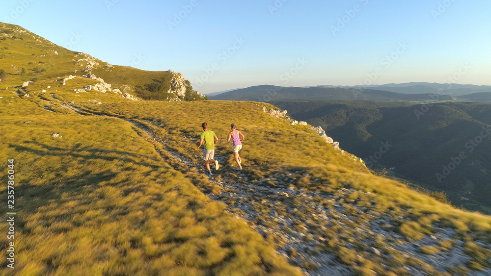 AERIAL: Young couple runs up a grassy hill overlooking the picturesque valley.