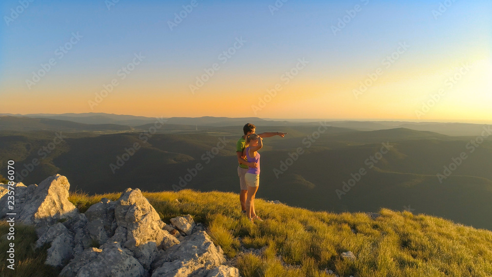AERIAL: Embraced man and woman observing the picturesque mountains at sunset.
