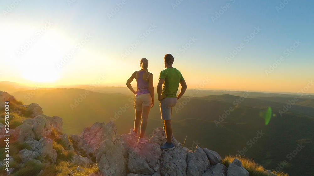 Fototapeta premium AERIAL: Flying near hiker couple watching the sunset after reaching the summit.