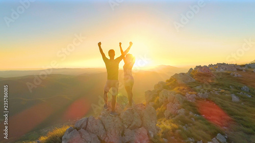 AERIAL: Hiker couple outstretches arms after reaching the summit at sunset.