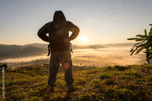 Silhouette of happy tourist man with backpack standing on top mountain looks into the distance at morning sunrise with foggy mountain landscape