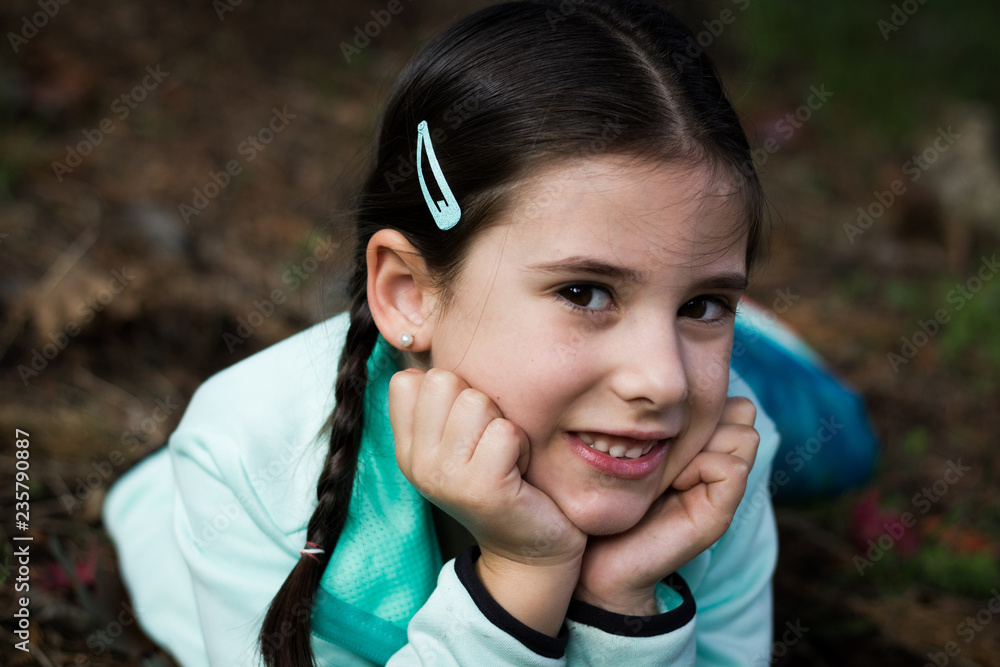 Lovely brunette girl portrait with braids resting in the park. Closeup of pretty young kid with hands on face looking at camera, positive thinking concepts