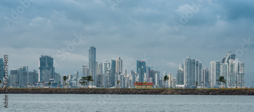 Hot, humid day in Panama city as another rainstorm brews quickly over the city skyline.  Tall buildings shimmer in heatwaves rising in humid air.  People on Panama Canal jetty park in foreground. © valleyboi63