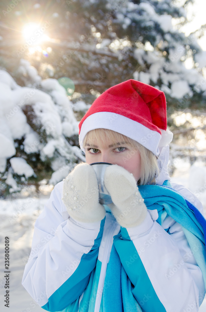 Obraz premium beautiful blond girl in a red Christmas hat is drinking hot tea from a cup. vertical view