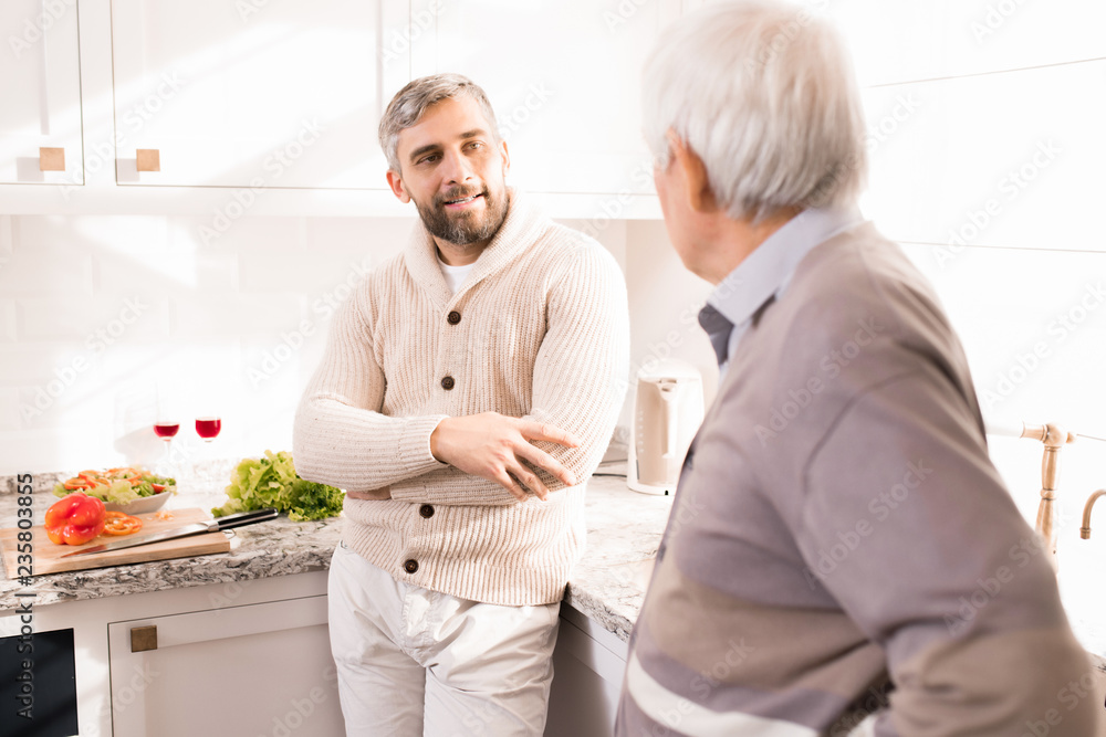 Obraz premium Warm toned portrait of bearded adult man talking to his father in kitchen lit by sunlight