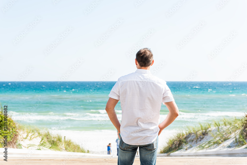 Back of young man standing by beach shore at sunny day in Seaside, Florida panhandle town village with ocean, wooden boardwalk, white shirt, jeans