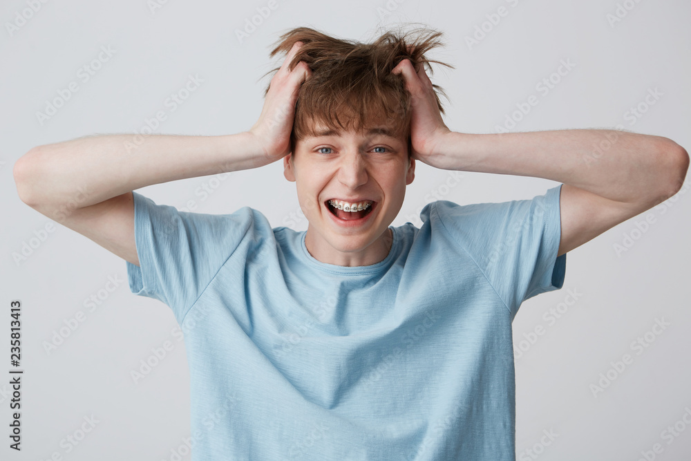 Screaming amazed excited young man clutched at his head, hair tousled, mouth wide opened as shouting loud with braces on teeth wears blue t-shirt, feels happy surprised isolated over white background Screaming amazed excited young man clutched at his head, hair tousled, mouth wide opened as shouting loud with braces on teeth wears blue t-shirt, feels happy surprised isolated over white background