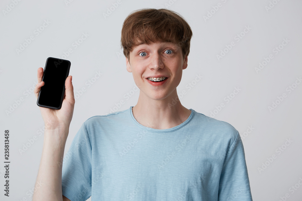 Young guy standing looking straight joyful smiling holding phone in his hand which is turned by a black screen to the camera. Portrays the joy of a new phone, unexpectedly an amazing message has come Young guy standing looking straight joyful smiling holding phone in his hand which is turned by a black screen to the camera. Portrays the joy of a new phone, unexpectedly an amazing message has come
