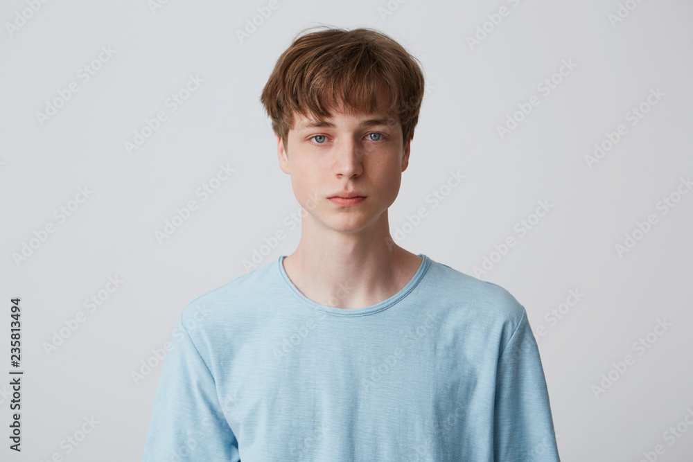 Close up face of a young man without emotions. Beautiful emotionless guy in a blue t-shirt looking to the camera, isolated over white background - obrazy, fototapety, plakaty 