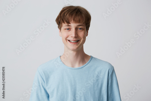 Teenager with short hair and braces on teeth looks camera, wears blue t-shirt, feels happy glad, smiling isolated over white background