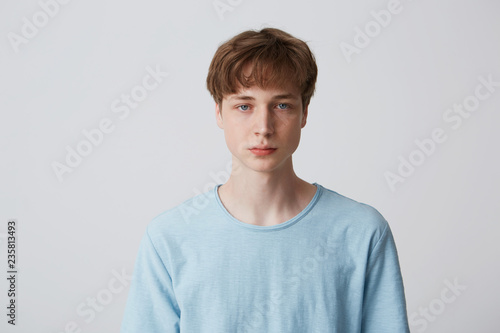 Headshot of a blue-eyed guy without emotions. Emotionless teenager in a blue t-shirt looking to the camera, isolated over white background photo