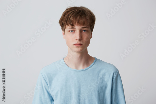 Close up face of a young man without emotions. Beautiful emotionless guy in a blue t-shirt looking to the camera, isolated over white background photo