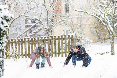 Young man, woman playing, sitting, making snowballs in winter snowstorm, storm, snowing at home, house garden, front yard, backyard, trees covered in snow, smiling, happy, laughing © Andriy Blokhin