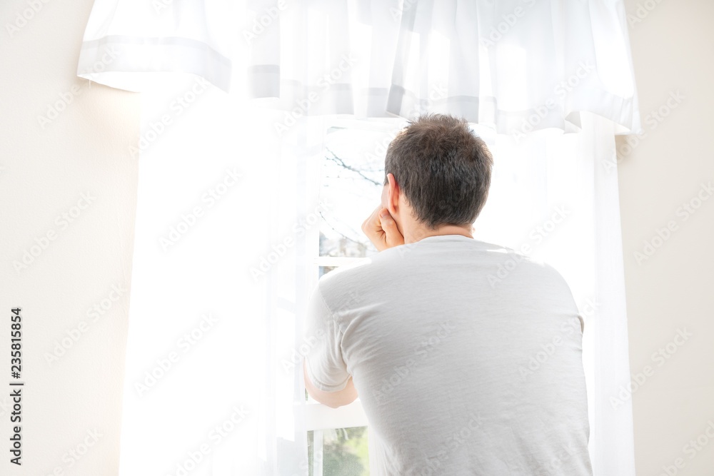 Back of one young male, man, standing, leaning arms, hands against head, looking out, through in white, bright room of house, home, apartment, sunny large window with blinds, curtains, wall
