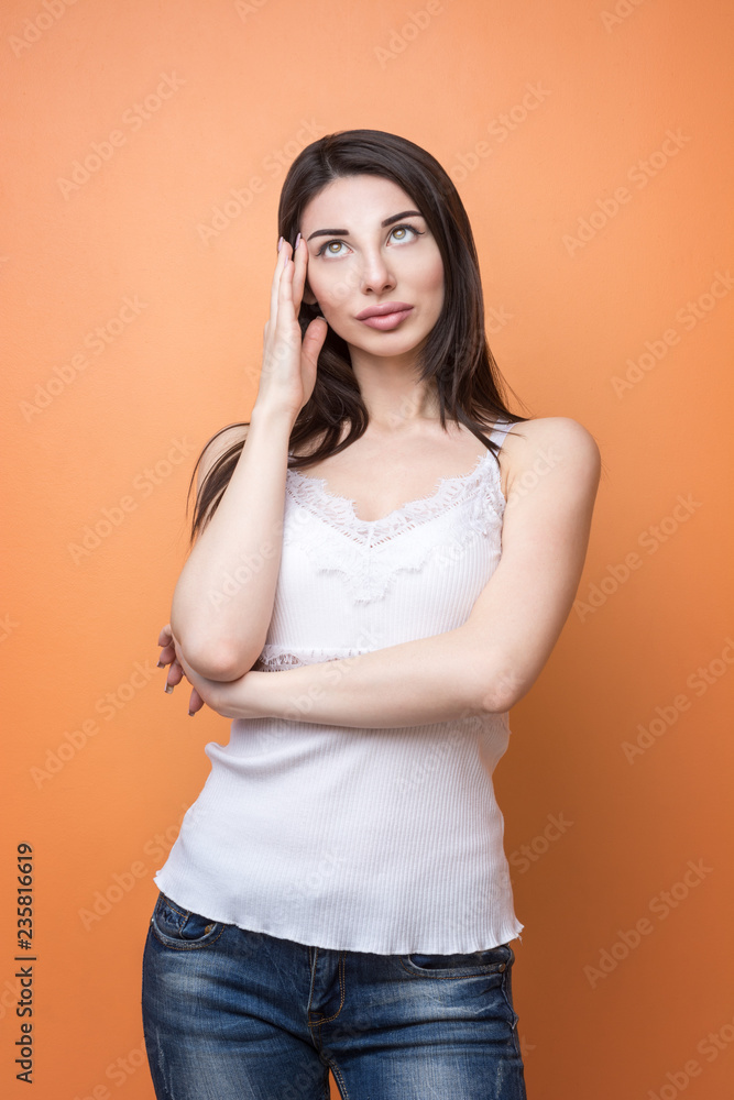 Closeup portrait of a young beautiful brunette pensive woman holding her hand new her head and looking up, think about something against orange background Closeup portrait of a young beautiful brunette pensive woman holding her hand new her head and looking up, think about something against orange background