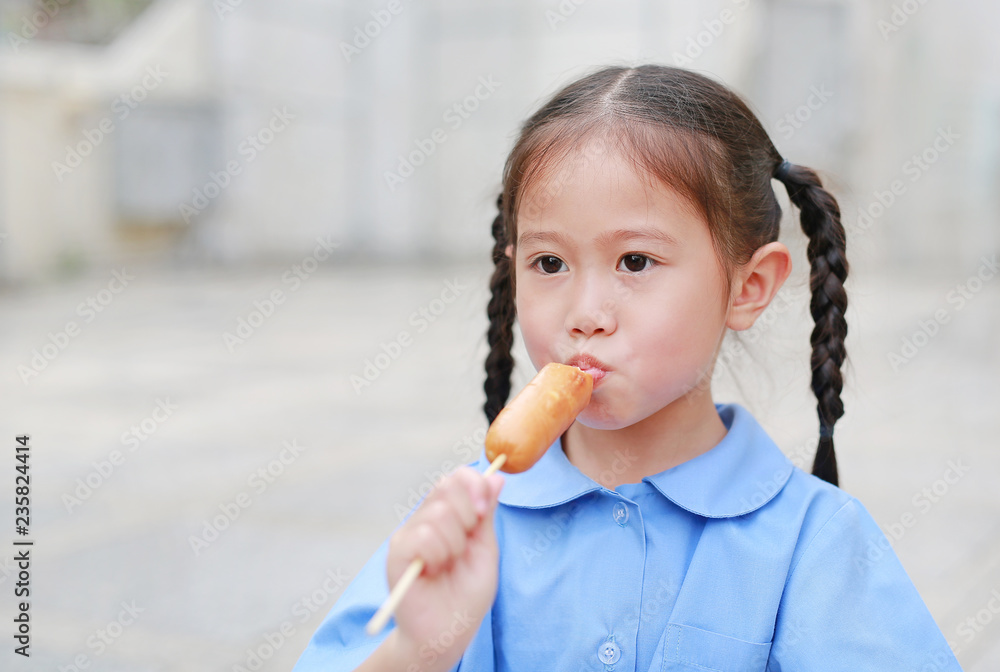 Adorable little Asian child girl in school uniform enjoy eating sausage. Adorable little Asian child girl in school uniform enjoy eating sausage.