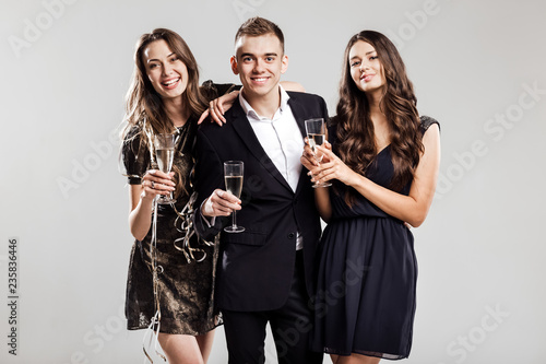 Party time. Two beautiful girls dressed in stylish elegant dresses and handsome man in the whit shirt smile and hold glasses of champagne