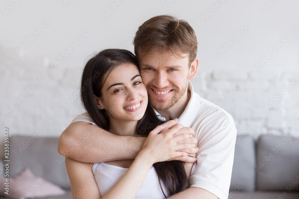 Head shot portrait of loving embracing happy couple in living room, smiling boyfriend standing behind attractive girlfriend, hugging, man and woman holding hands, family portrait, look at camera