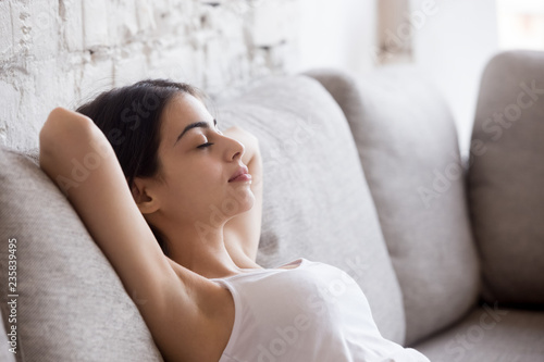Young calm woman relaxing leaning back with hands behind head on comfortable sofa, having daydream, tired girl sleeping, napping on couch in living room, breathing calm, no stress free weekend at home