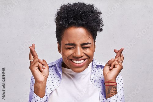 Photo Beautiful African American female student crosses fingers with big hope, has positive expression, poses against the blank background