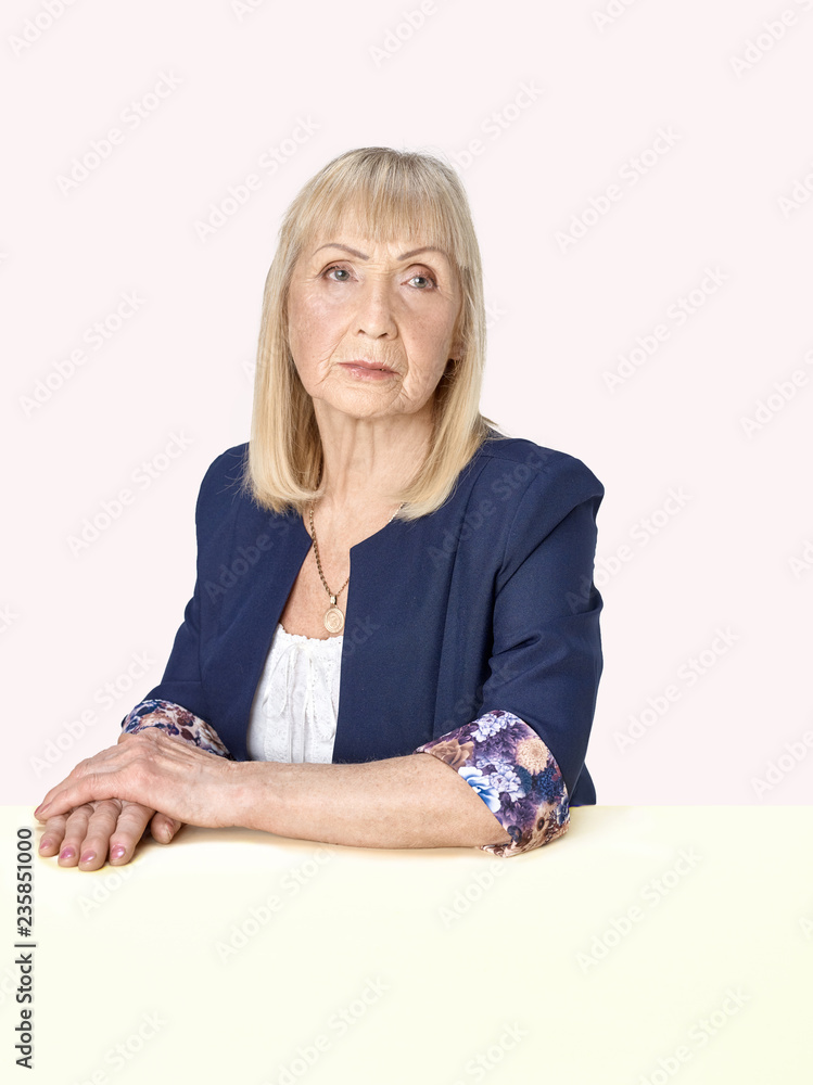 Senior woman sitting at table at studio and thinking about her sad life against pastel background. Human emotions and facial expression concept Senior woman sitting at table at studio and thinking about her sad life against pastel background. Human emotions and facial expression concept