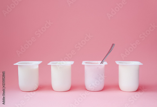 Four yogurts in white plastic cups on pink background with copy space. Strawberry pink yoghurt with spoon in it. Minimal style. Concept of better choice.