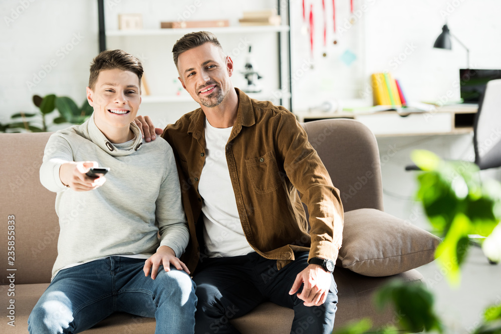 smiling father and teen son watching tv together on sofa in living room smiling father and teen son watching tv together on sofa in living room