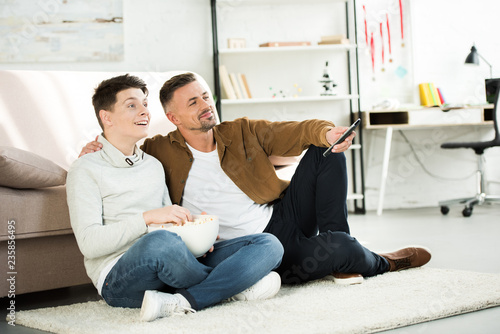 happy father and teen son watching tv and holding bowl of popcorn on floor at home