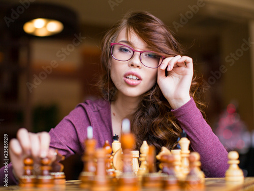 Close up of woman in glasses playing chess game. Looks at camera.