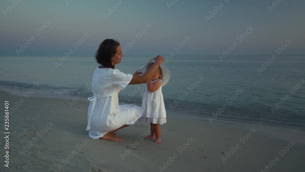 Young beautiful woman walks with her little daughter on the seashore at sunset. Mom and her cute little daughter try on a white hat. They are happy. Slow motion. Young beautiful woman walks with her little daughter on the seashore at sunset. Mom and her cute little daughter try on a white hat. They are happy. Slow motion.